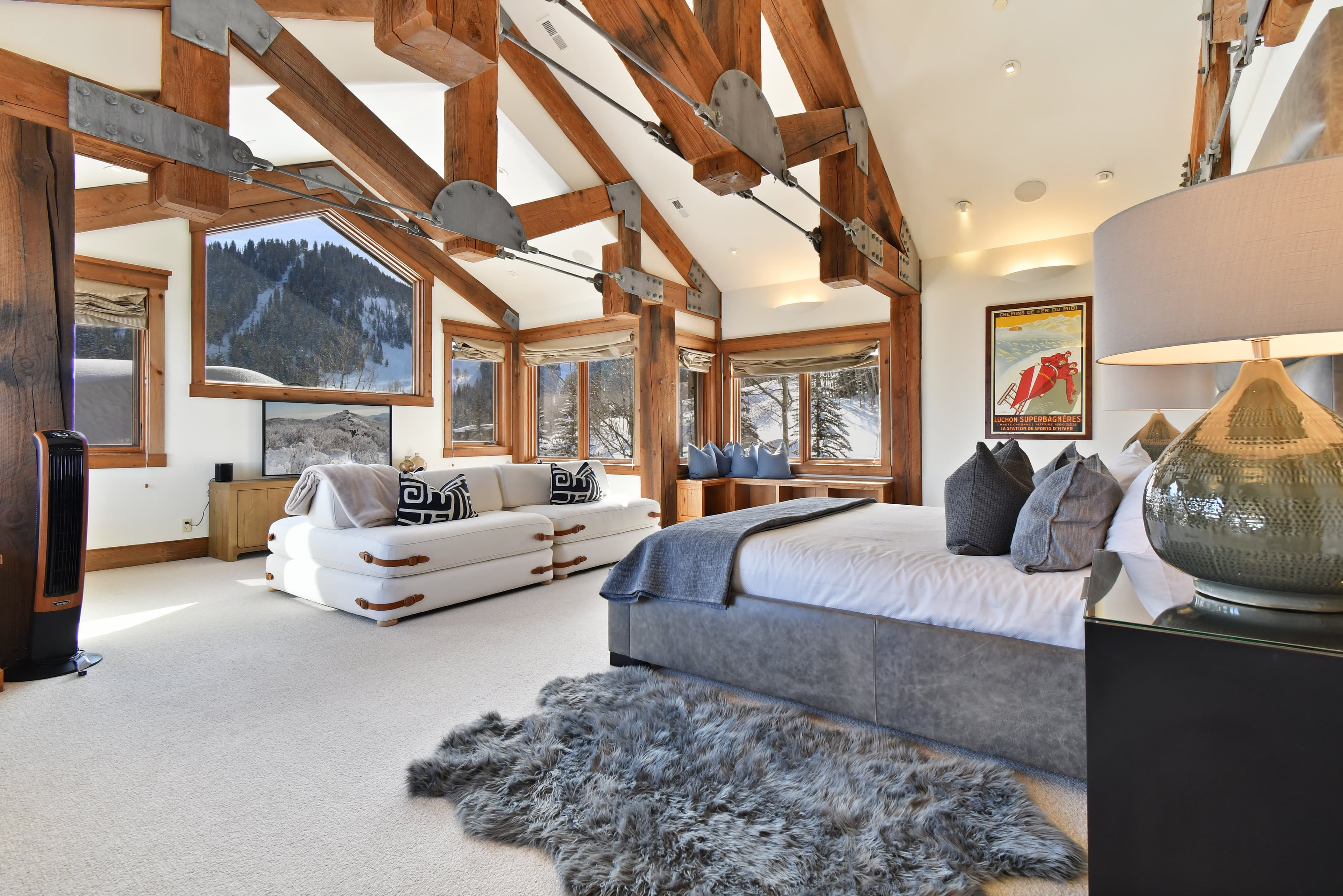 Large windows sit in front of the bed of the primary bedroom, with a large couch in front of an entertainment center and TV.