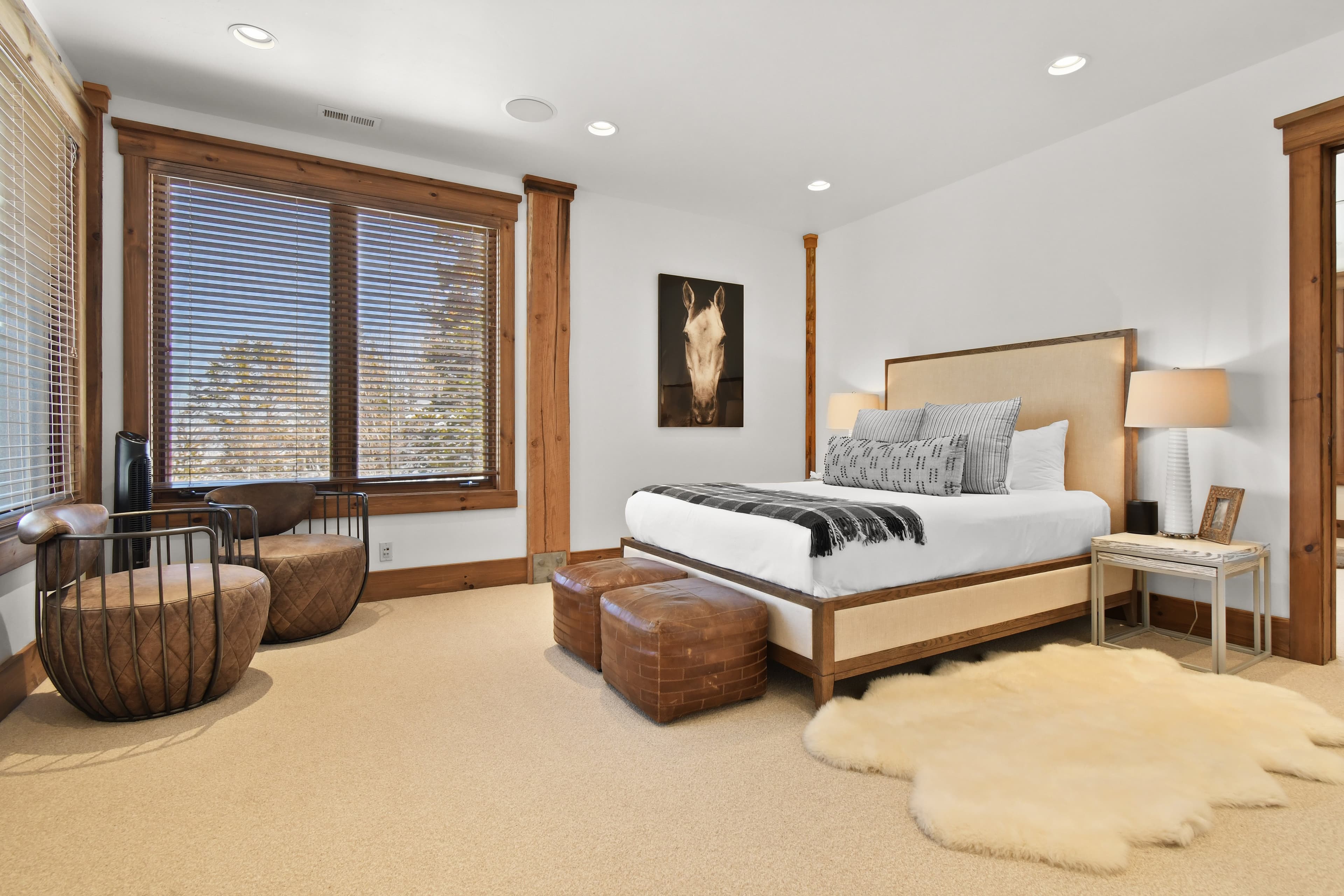A white fur rug is set beside a large bed, with two leather ottomans at the foot of it.